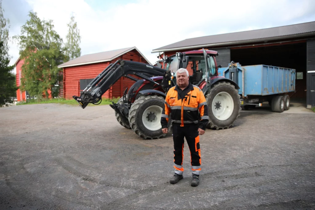 Viljanviljelijä ja lumiurakoitsija Juha Tamminen seisoo punaisen traktorinsa edessä. Traktorin sininen peräkärry on puoliksi takana olevan tallin sisällä. Taustalla näkyy myös kaksi punaista puurakennusta ja muutama koivu.